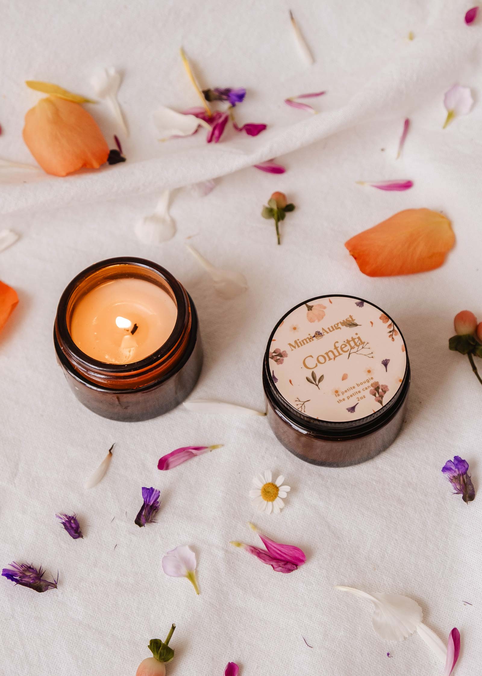 Une bougie allumée Mimi & August Confetti 2oz - Bougie réutilisable dans un pot brun aux notes subtiles de mandarine est posée sur un tissu au milieu de pétales de fleurs colorées. À proximité, un autre pot avec un couvercle floral renforce l'ambiance du parfum fruité.