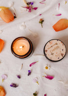 The Confetti - Reusable Candle by Mimi & August, encased in a brown jar with a floral lid, sits on a white cloth surrounded by flower petals and small blooms, emitting delightful mandarin notes that blend seamlessly with the setting.