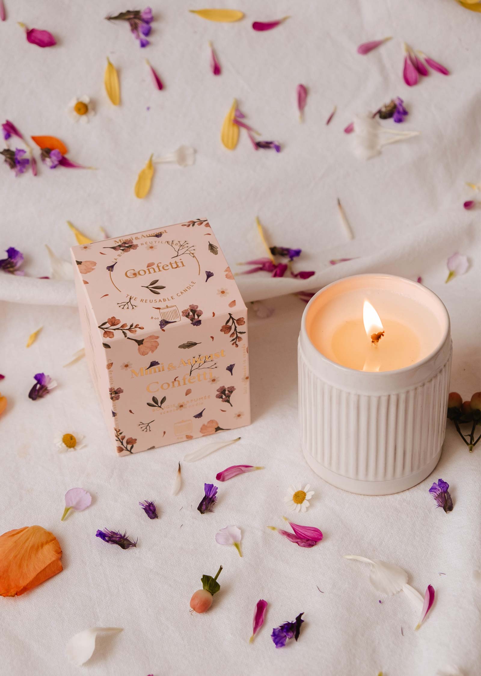 A lit Mimi & August's Confetti - Reusable Candle in a ceramic holder rests on white fabric, surrounded by scattered flower petals and a decorative box. Its subtle mandarin scent enhances the serene setting with a fruity aroma.