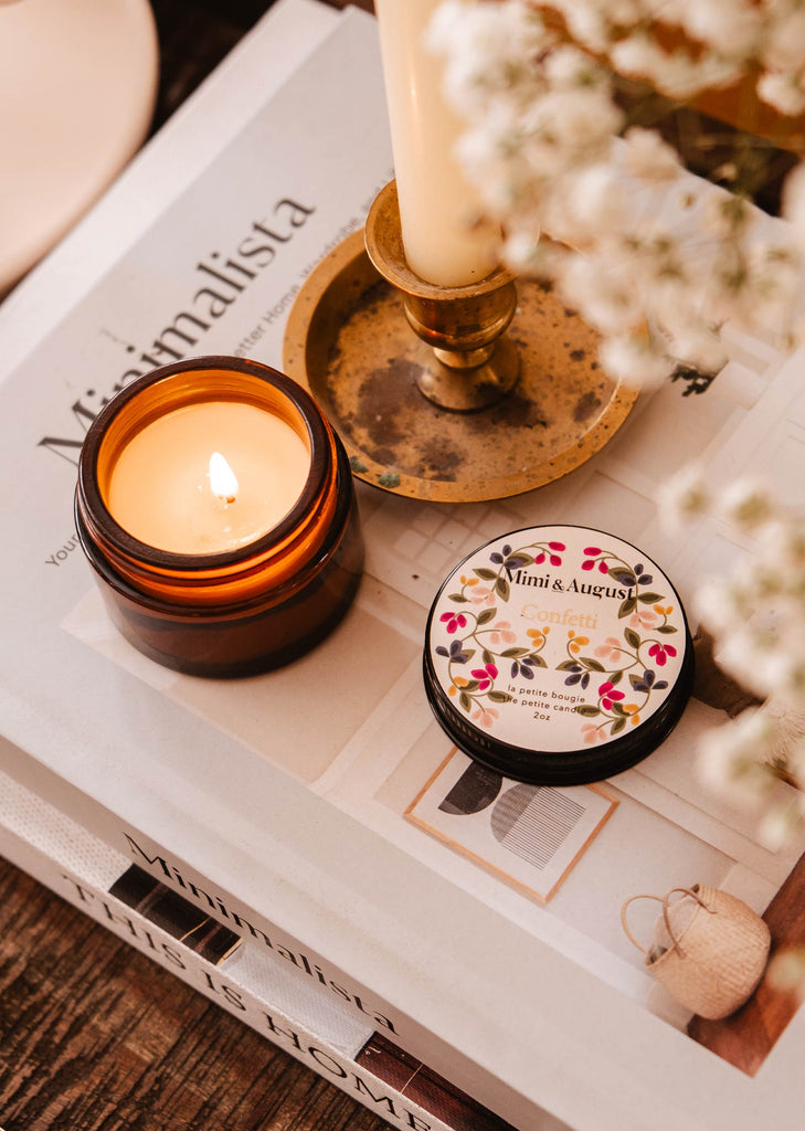 A Confetti - Reusable Candle from Mimi & August sits on a table next to a book, adding a touch of celebration to the ambiance.