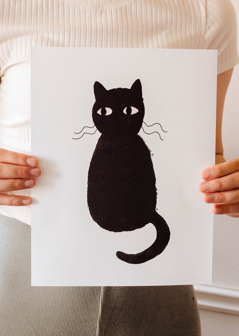 A woman displaying a Cute Black Cat Art Print by Mimi & August on recycled paper.