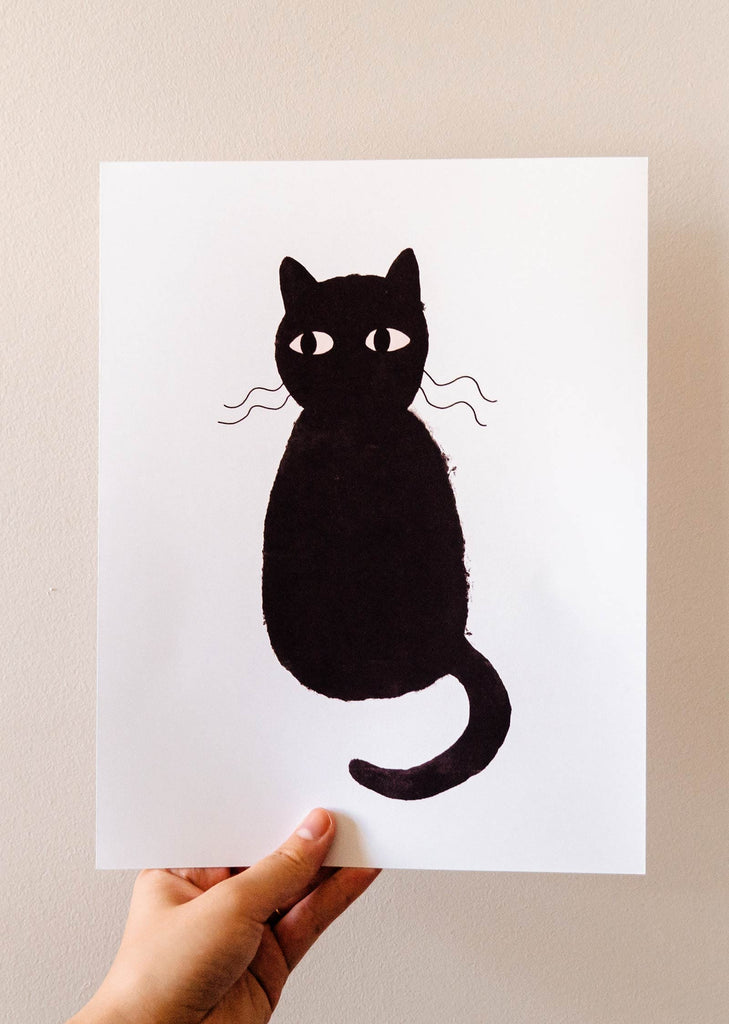 A person showcasing a Cute Black Cat Art Print by Mimi & August on recycled paper.