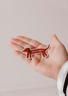 A hand holds the "Dachshund Dog - Vinyl Sticker" by Mimi & August, a durable, weatherproof vinyl sticker shaped like a brown dachshund with a minimalist design and the brand's name on its body.