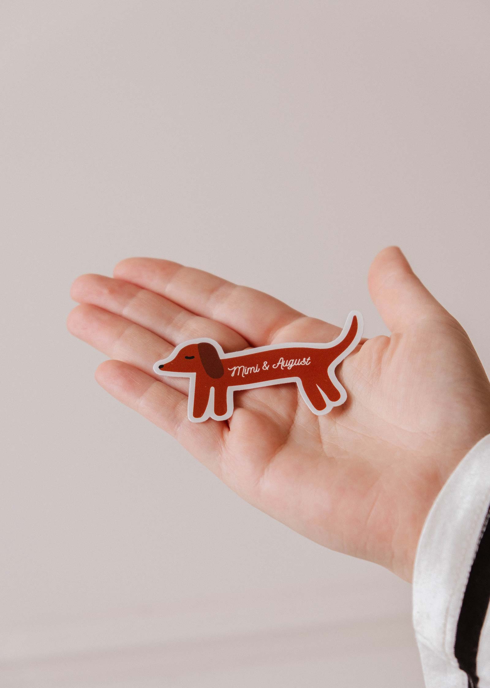 A hand holds the "Dachshund Dog - Vinyl Sticker" by Mimi & August, a durable, weatherproof vinyl sticker shaped like a brown dachshund with a minimalist design and the brand's name on its body.