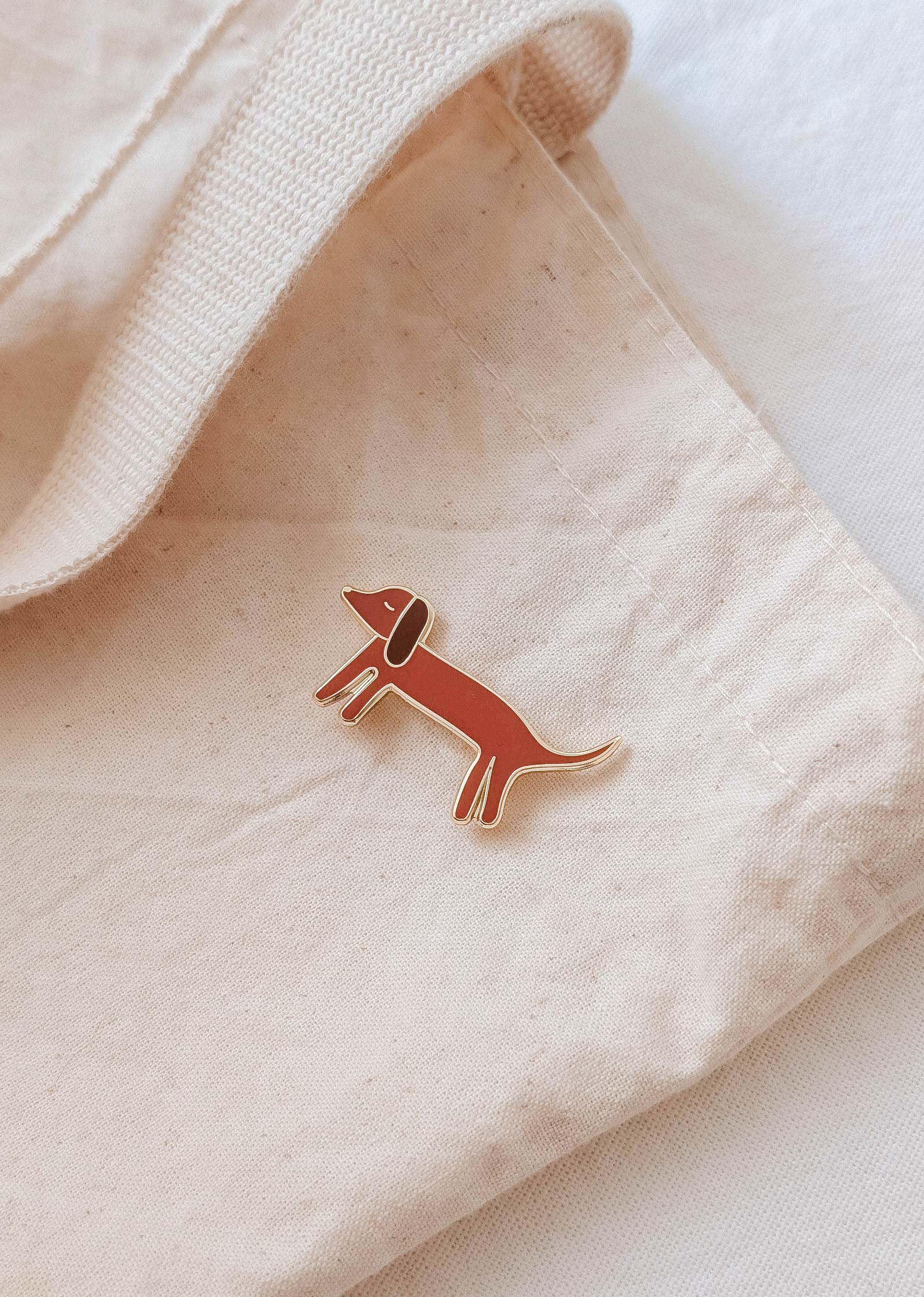 The Mimi & August Dachshund Enamel Pin, shaped like a wiener dog, is attached to a beige fabric tote bag—an adorable accessory for any dog lover.