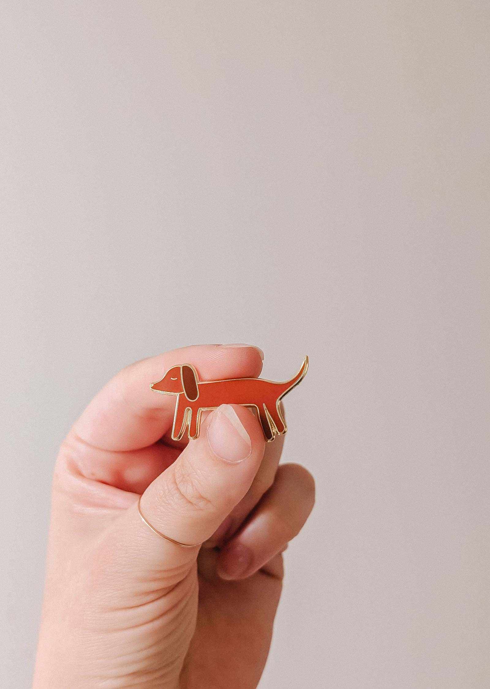 A hand holds the Mimi & August Dachshund Enamel Pin, shaped like a brown wiener dog—perfect for dog lovers—against a plain light background.