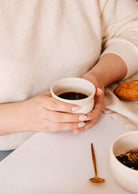 Une personne tient une tasse Douce Matin - Ceramic Cup by Mimi & August, ainsi qu'une cuillère en or, un bol de céréales et une pâtisserie à table.