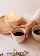 A hand gently cradles the Douce Matin ceramic cup from Mimi & August, amidst a lit candle and dried fruit bowl, creating an inviting scene.