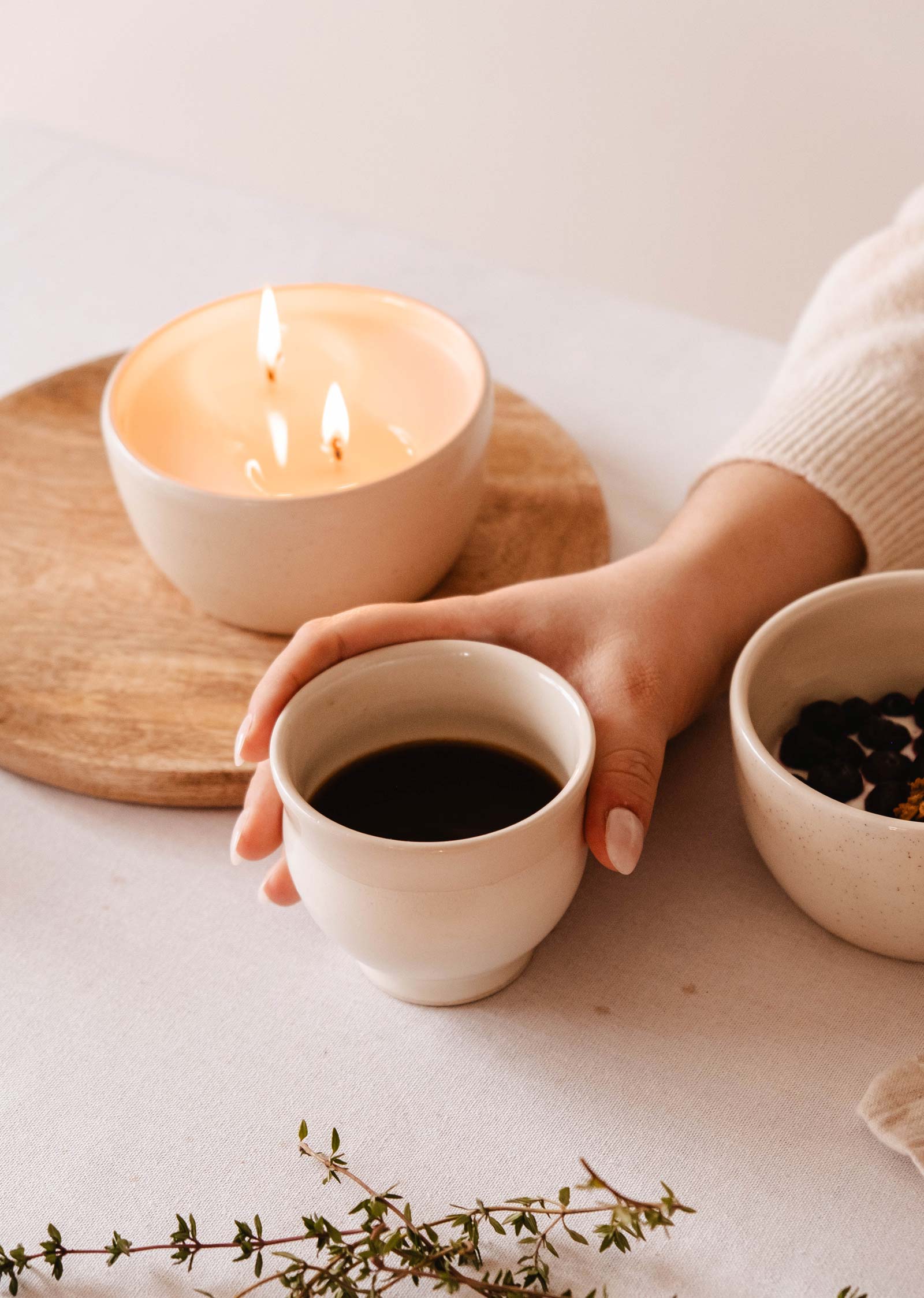 A hand gently cradles the Douce Matin ceramic cup from Mimi & August, amidst a lit candle and dried fruit bowl, creating an inviting scene.