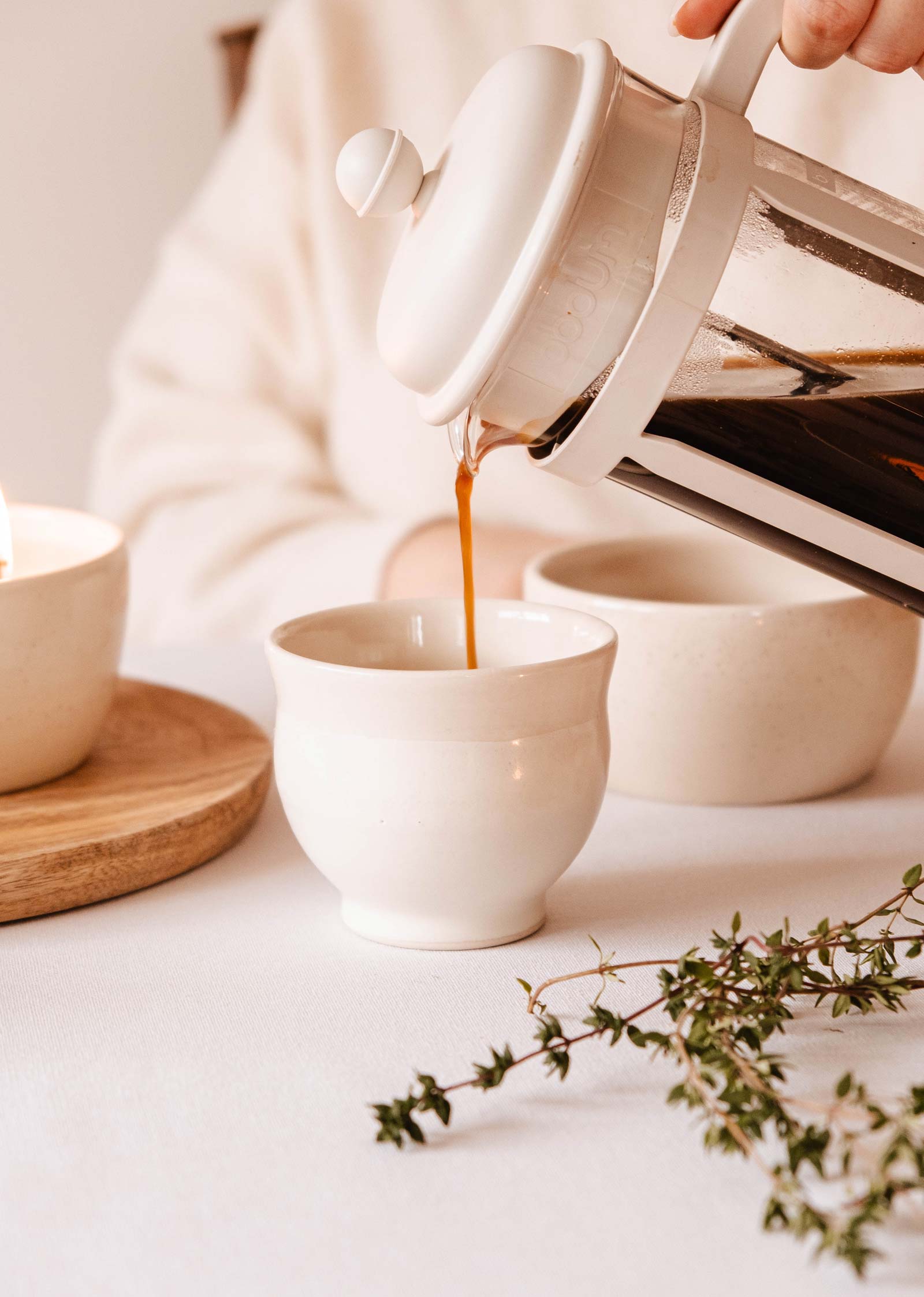 Le café est versé d'une presse française dans la tasse en céramique Douce Matin - Ceramic Cup de Mimi & August sur une table avec des bols blancs et des herbes vertes, soulignant la beauté de la vaisselle polyvalente fabriquée à la main.