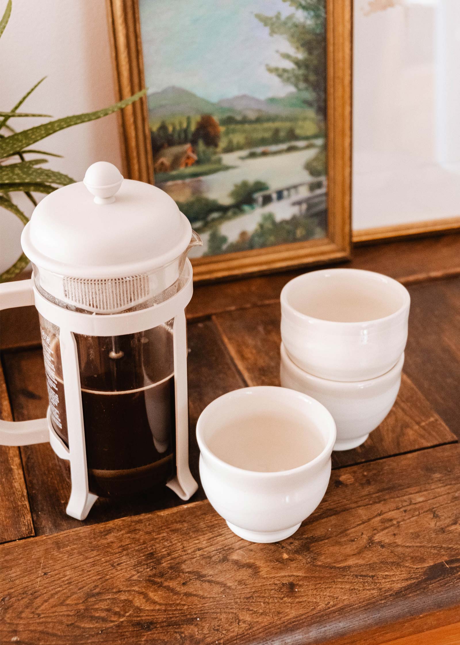 A white French press filled with coffee exudes artisanal charm on a wooden table, paired with three stacked Douce Matin - Ceramic Cups by Mimi & August. In the background, a framed landscape painting adds elegance to the handcrafted scene.