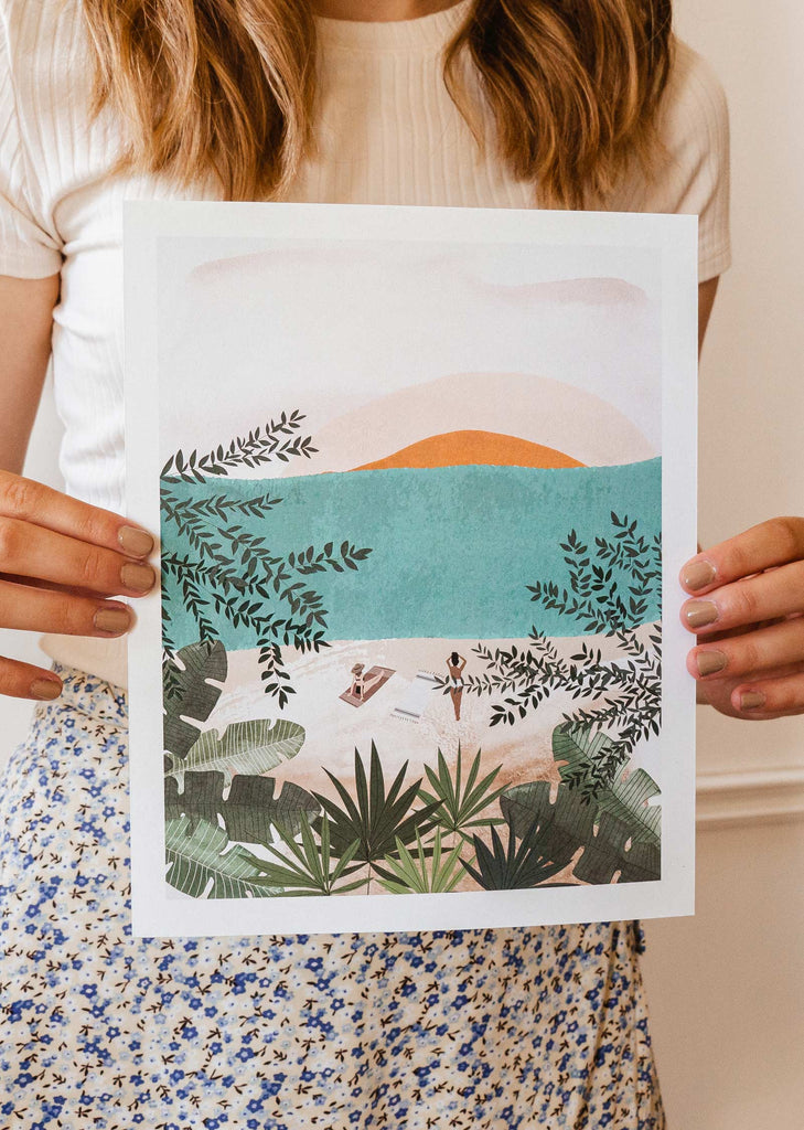A woman holding up an art print of a mimi and august tropical beach scene.