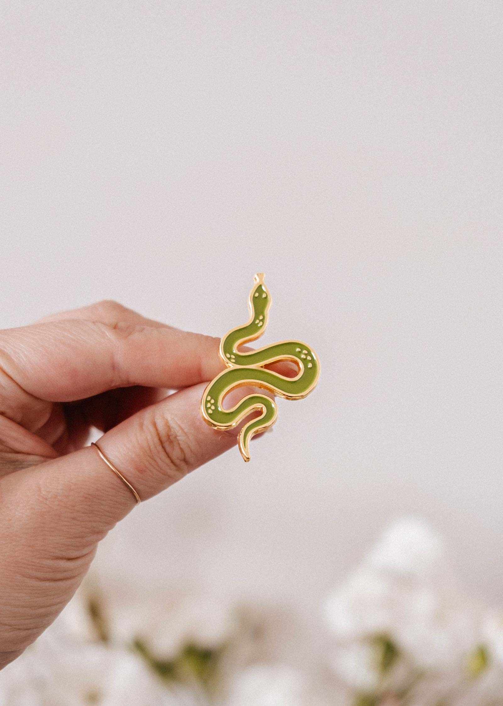 A hand displays the Mimi & August Cute Serpent Enamel Pin, shaped like a green and yellow snake, against a blurred light background—perfect for animal lovers.