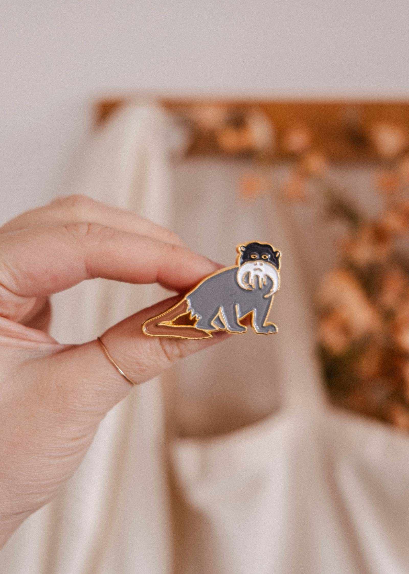 A hand displays the Sir Moustache Enamel Pin by Mimi & August—a charming gray and white monkey with a curled tail—against a softly blurred beige and brown background, ideal for animal lovers.