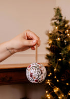 A hand delicately holds an Enchanted Forest - Christmas Ornament Candle from Mimi & August, glistening enchantingly in front of a lit Christmas tree.