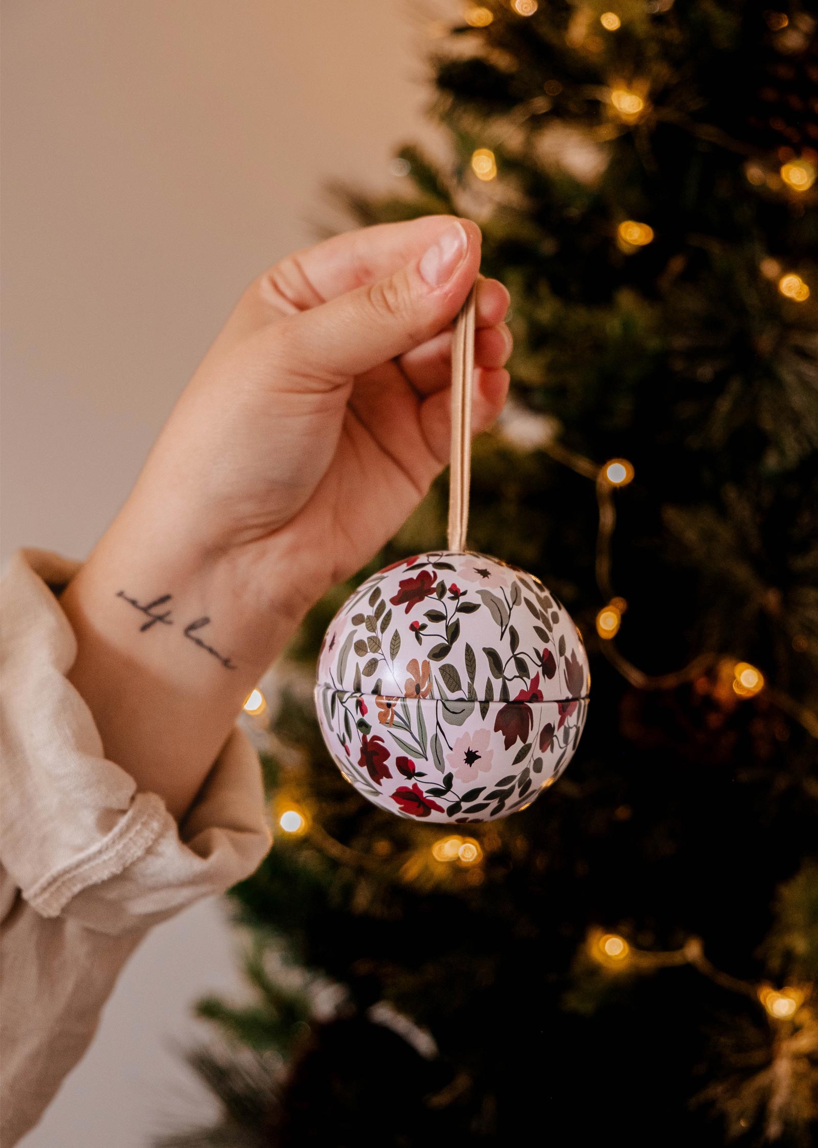 Une main tient une bougie Enchanted Forest - Christmas Ornament Candle by Mimi & August devant un arbre de Noël sur le thème de la forêt enchantée, orné de lumières scintillantes.