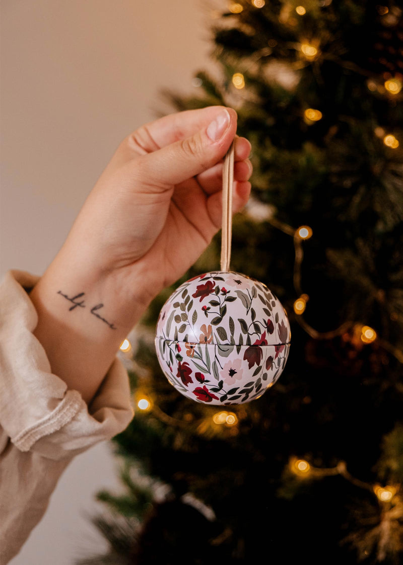 A hand holds an Enchanted Forest - Christmas Ornament Candle by Mimi & August in front of an enchanted forest-themed Christmas tree adorned with twinkling lights.