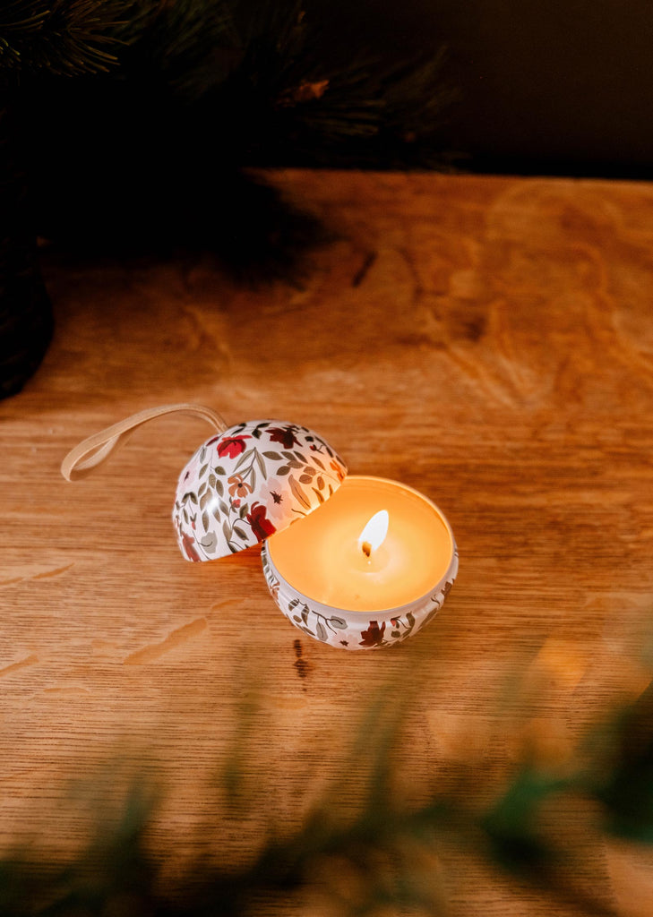 The Enchanted Forest - Christmas Ornament Candle by Mimi & August rests elegantly in a patterned holder on a wooden surface, surrounded by greenery and Christmas baubles that partially appear in the foreground, creating an ambiance reminiscent of an enchanted forest.