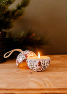 The Enchanted Forest - Christmas Ornament Candle by Mimi & August, nestled in a decorative tin adorned with floral patterns, adds an enchanting forest ambiance to a wooden surface, with its open lid placed nearby.