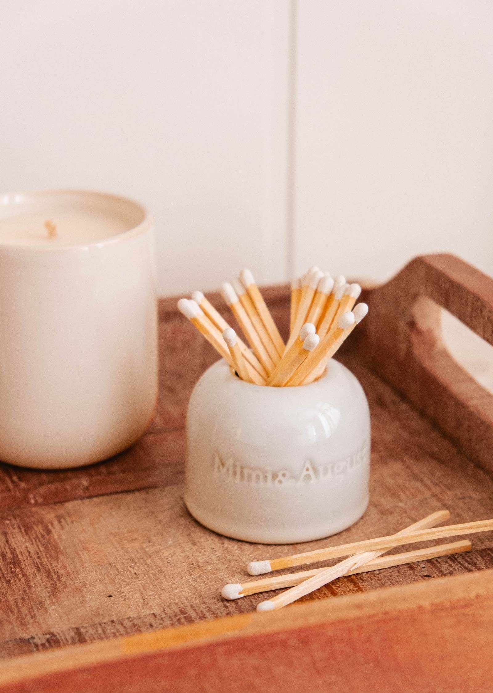 A white jar named "Étincelles – Ceramic Match Holder" by Mimi & August serves as a versatile holder for cotton swabs on a wooden tray, with a candle glowing softly nearby.