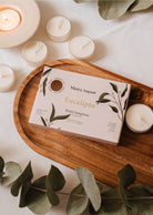 The Eucalipto Tealight Candle Set by Mimi & August is displayed on a wooden tray, surrounded by lit candles and eucalyptus leaves, creating a soothing atmosphere.