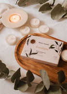 The Eucalipto Tealight Candle Set by Mimi & August rests on a wooden tray adorned with eucalyptus leaves and lit candles, creating a soothing spa ambiance on a white surface.