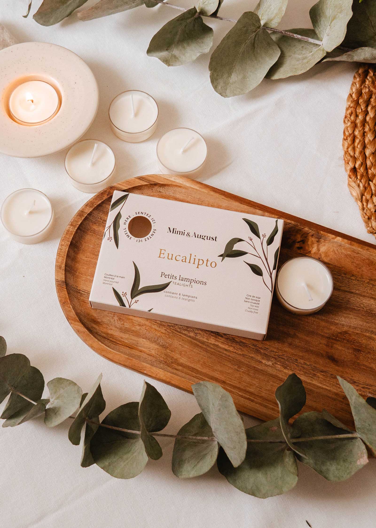 The "Eucalipto Tealight Candle Set" by Mimi & August rests on a wooden tray with eucalyptus leaves and lit candles, evoking a spa-like atmosphere that invites relaxation.