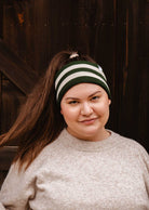 A person with long brown hair in a high ponytail, wearing the Mimi & August Evergreen Stripes Headband and a beige sweater featuring soft black fleece lining, stands in front of a dark wooden background.