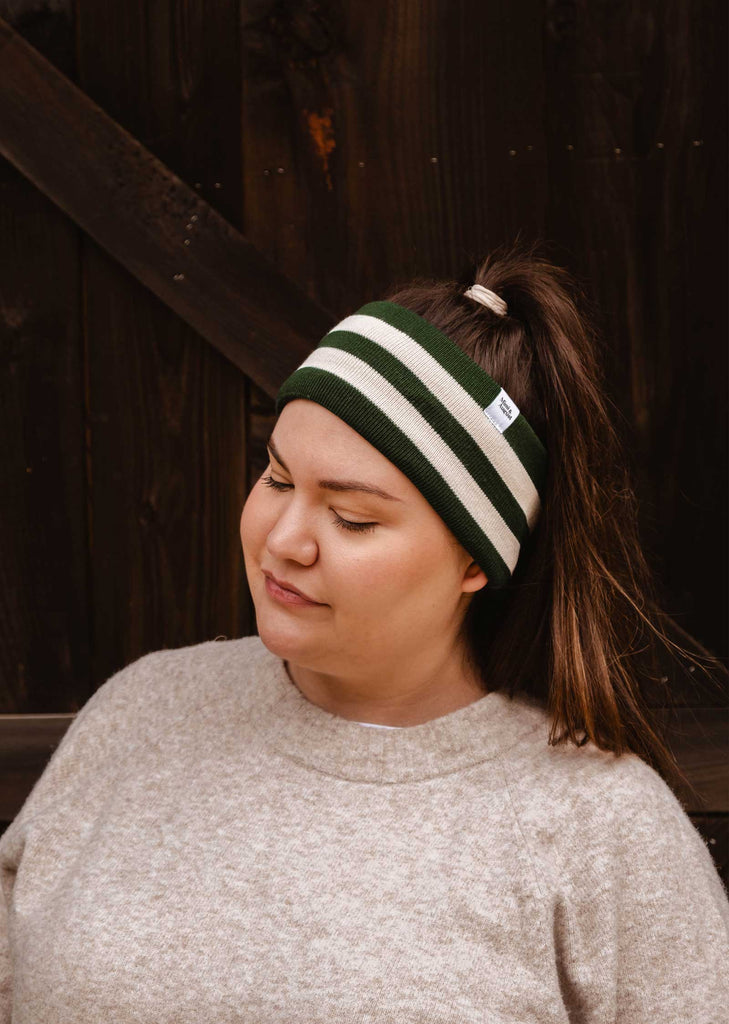 A person with long hair in a ponytail stands against a dark wooden background, wearing the Mimi & August Evergreen Stripes Headband—a green and white unisex acrylic knit with a cozy fleece lining—and a light grey sweater.