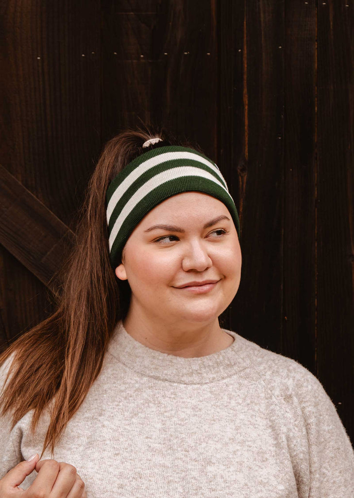 A person wearing a Mimi & August Evergreen Stripes Headband and a gray sweater looks to the side while standing in front of a dark wooden background. The unisex acrylic knit headband features green and white stripes, providing both style and warmth.