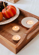 The Mimi & August Exclusive Discovery Kit with three lit candles on a wooden tray sits beside a plate displaying a decorative pumpkin and pinecones, all atop a white tablecloth.