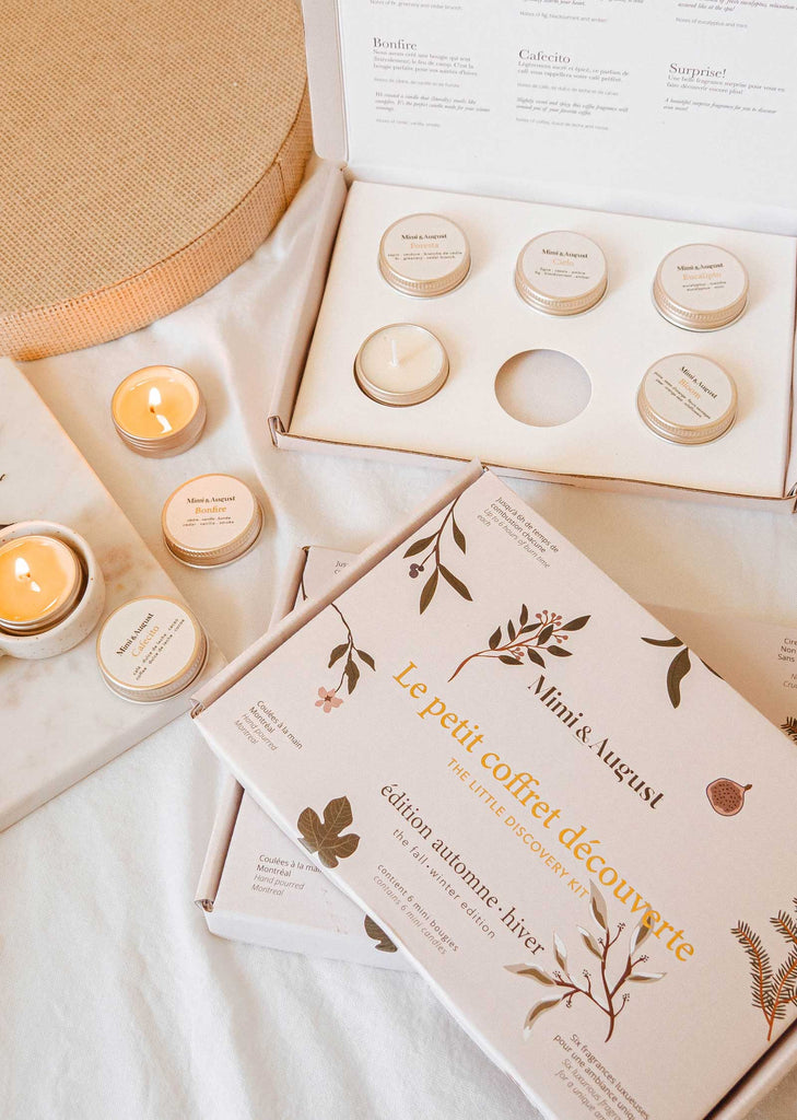 A collection of scented candles displayed in decorative boxes from Mimi & August. Some candles are lit, while others remain in their containers. The packaging features botanical illustrations and text in French, perfect for The Fall/Winter Discovery Kit full of cozy candle scents like the Foresta fragrance.