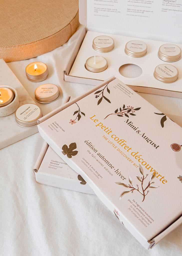 Close-up of a boxed set labeled "Le petit Coffret découverte" containing cozy candle scents and small tins on a light-colored surface with a lit candle nearby, part of Mimi & August's The Fall/Winter Discovery Kit.