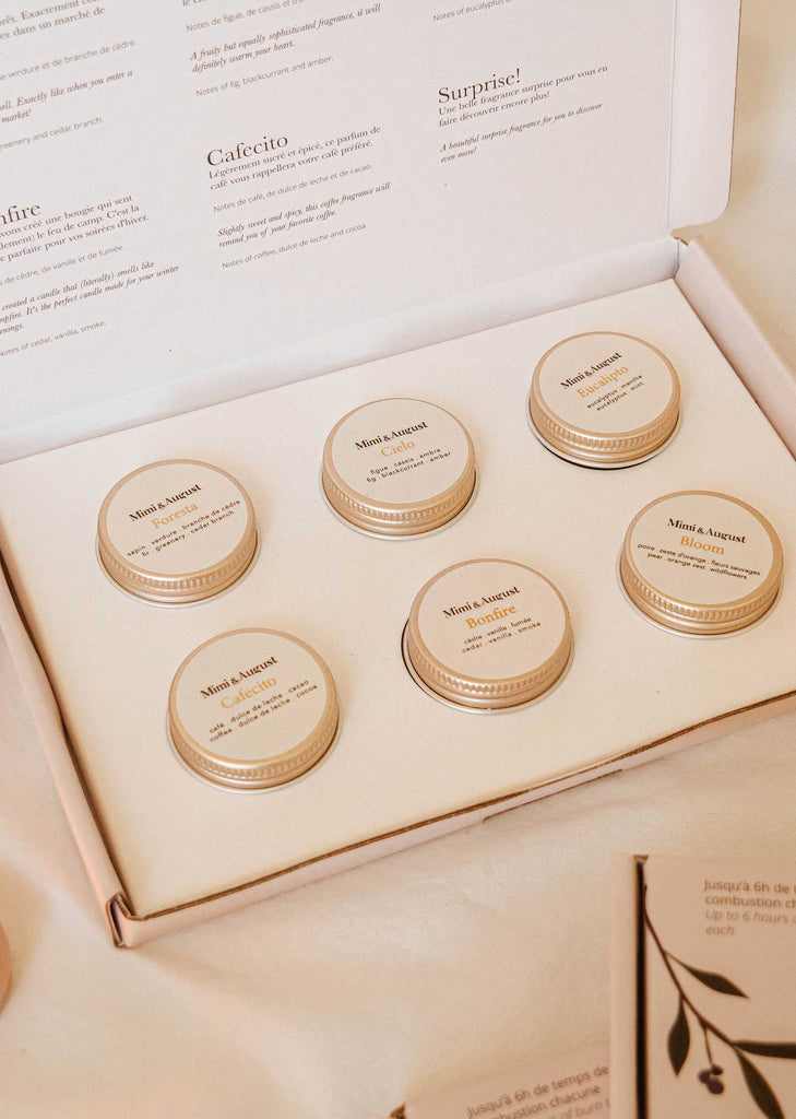 Open box containing six small jars labeled with cozy candle scents, displayed on a flat surface. Background includes a partially visible product information card from The Fall/Winter Discovery Kit by Mimi & August.