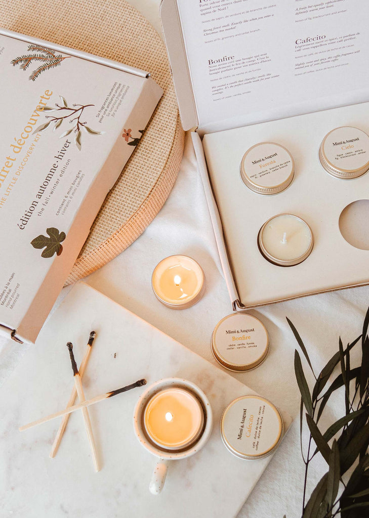 A collection of cozy candle scents on a white surface from Mimi & August, featuring a lit candle in a teacup, an open box with more candles, and matchsticks. The Fall/Winter Discovery Kit includes text printed on the inside of the box lid.