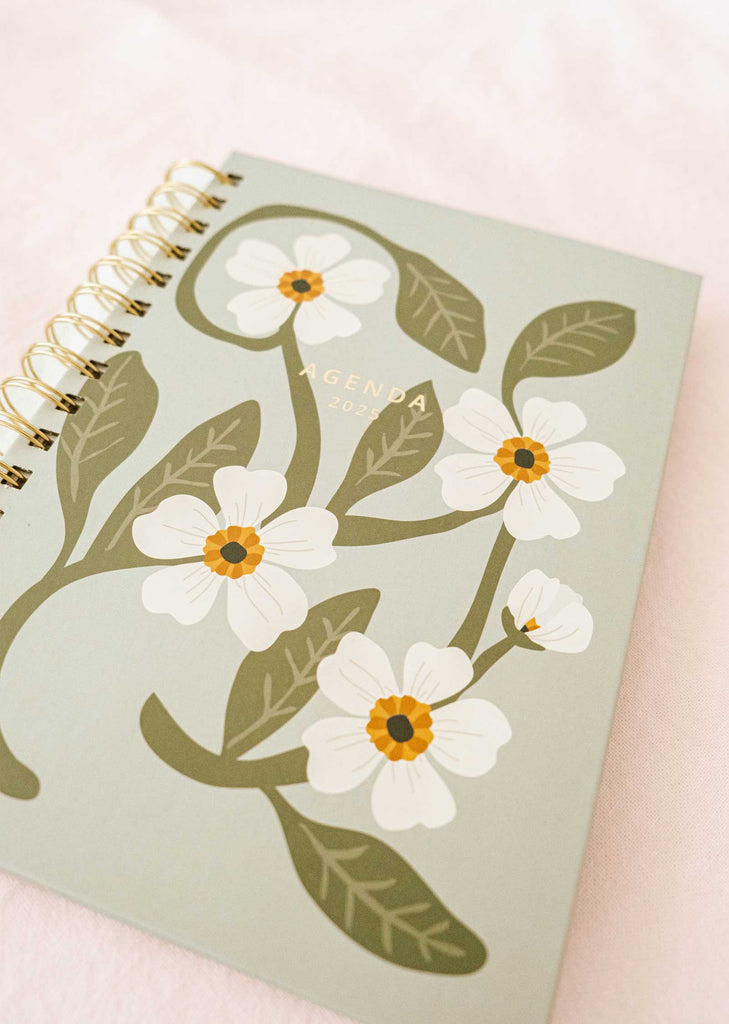 The Fields of Flowers - Agenda 2025 from Mimi & August features a durable cover adorned with white flowers and green leaves in a delightful floral design. Its robust spiral binding ensures it remains intact throughout the year.