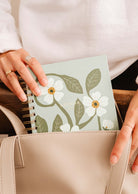 A person gently places the Fields of Flowers - Agenda 2025 by Mimi & August, featuring its durable cover, into a beige handbag.