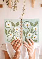 Someone holds a Fields of Flowers - Agenda 2025 by Mimi & August open, displaying its green pages decorated with white blossoms. Its robust spiral binding and durable cover make it perfect for enduring daily adventures.