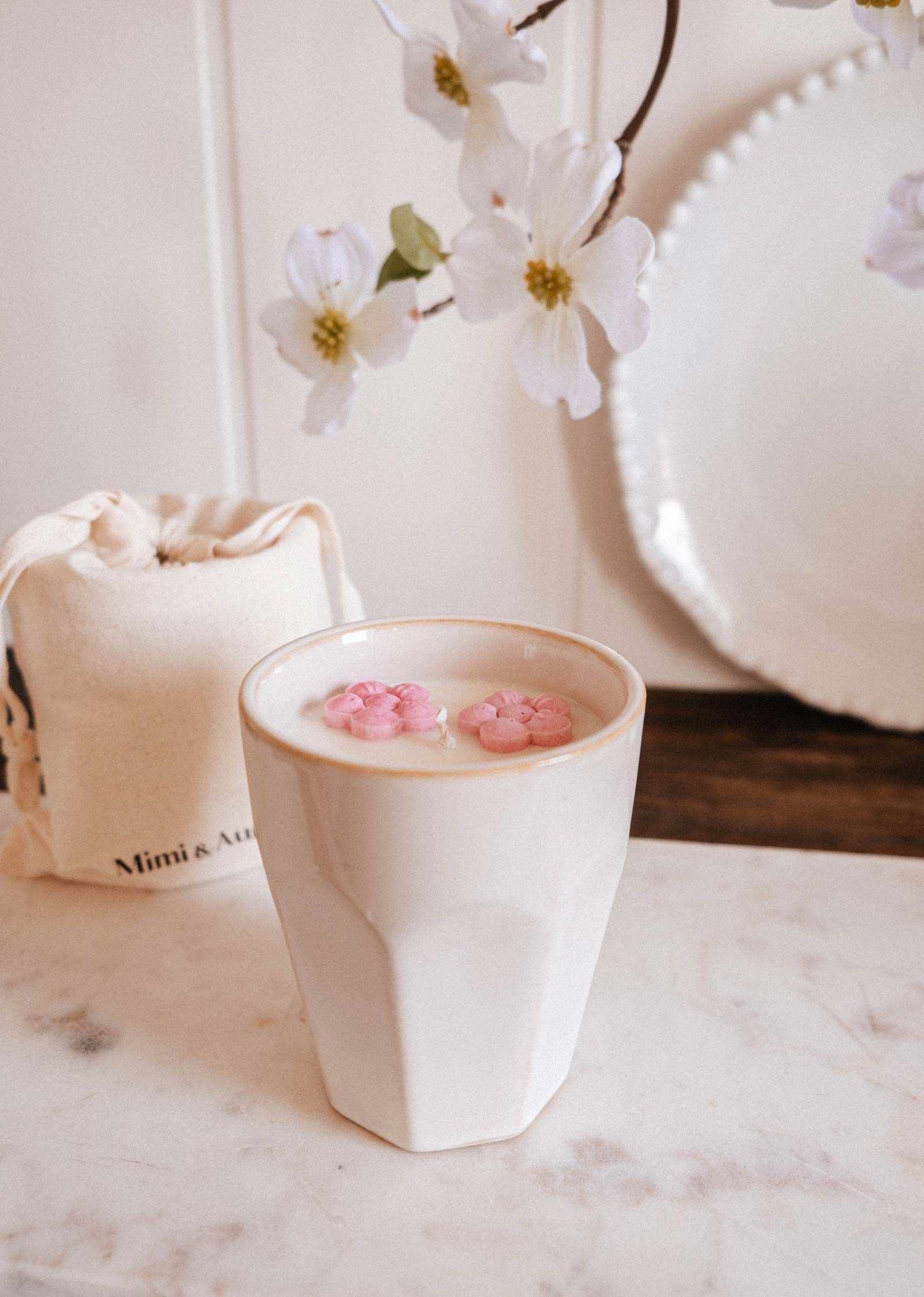 A white ceramic cup with a creamy drink and pink petals rests on marble, beside white flowers and a drawstring bag—perfect to pair with the Mimi & August Special Edition Candle for an added touch of elegance.