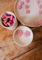 Pink flower-shaped candies on a ceramic plate, with more in a white cup and two floating in milk beside the Mimi & August Special Edition Candle on a wooden table.