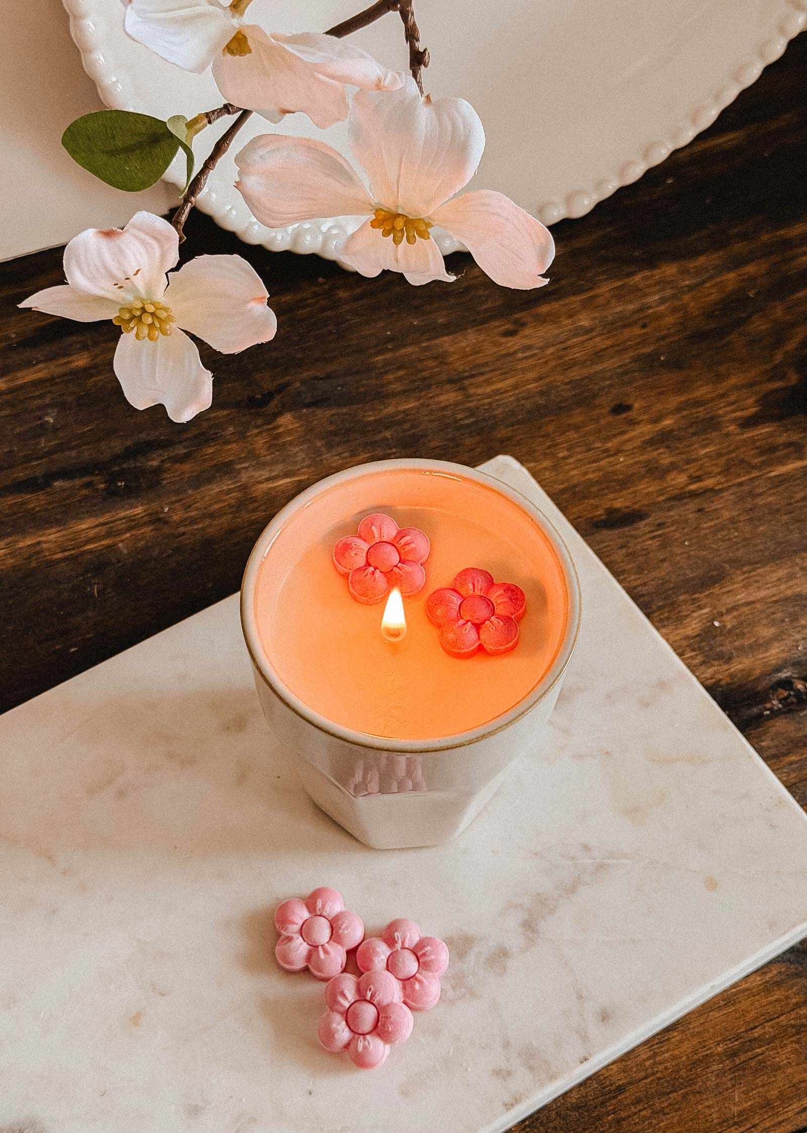 The Mimi & August Special Edition Candle, featuring two pink flower-shaped wax melts, is displayed on a marble slab beside artificial flowers and an extra pink flower-shaped wax melt.