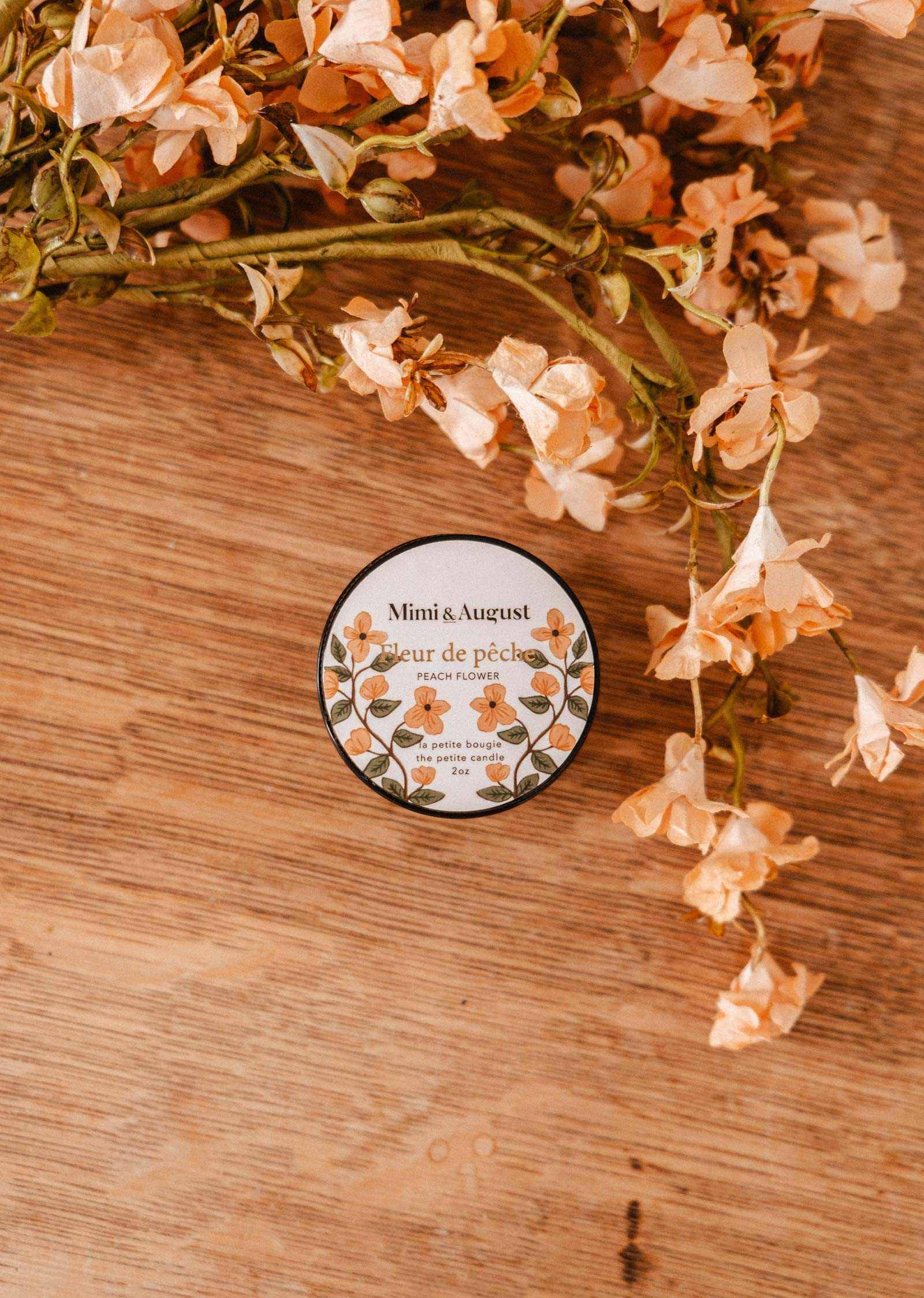A 2 oz Mimi & August Fleur de Pêche soy candle rests on a wooden surface beside peach-toned artificial flowers, highlighting the hand-poured floral charm of this Montreal-made candle.