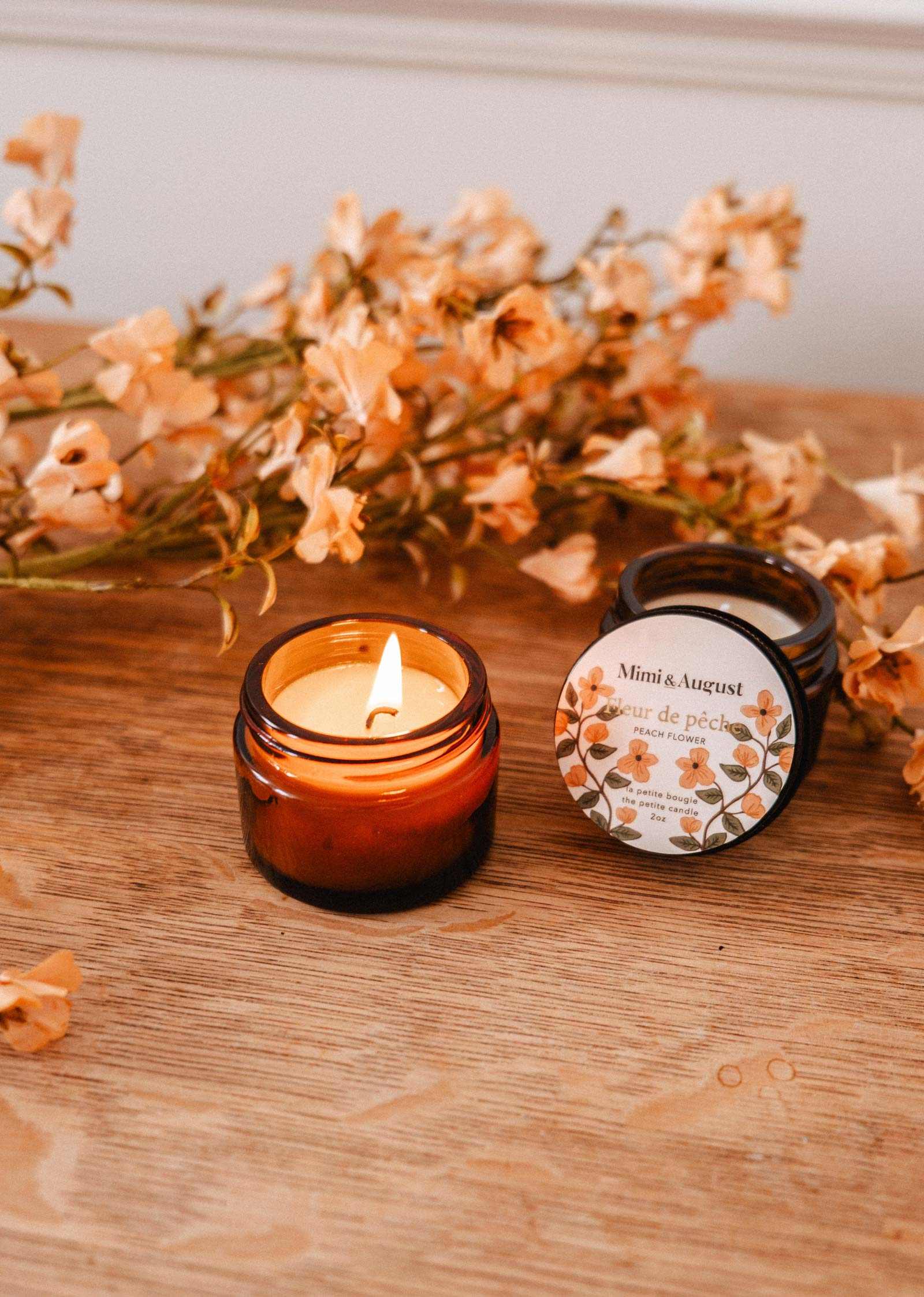 The Mimi & August Fleur de Pêche 2 oz soy candle, hand-poured in Montreal, glows in its amber glass jar on a wooden surface beside its lid, with peach-colored flowers in the background.