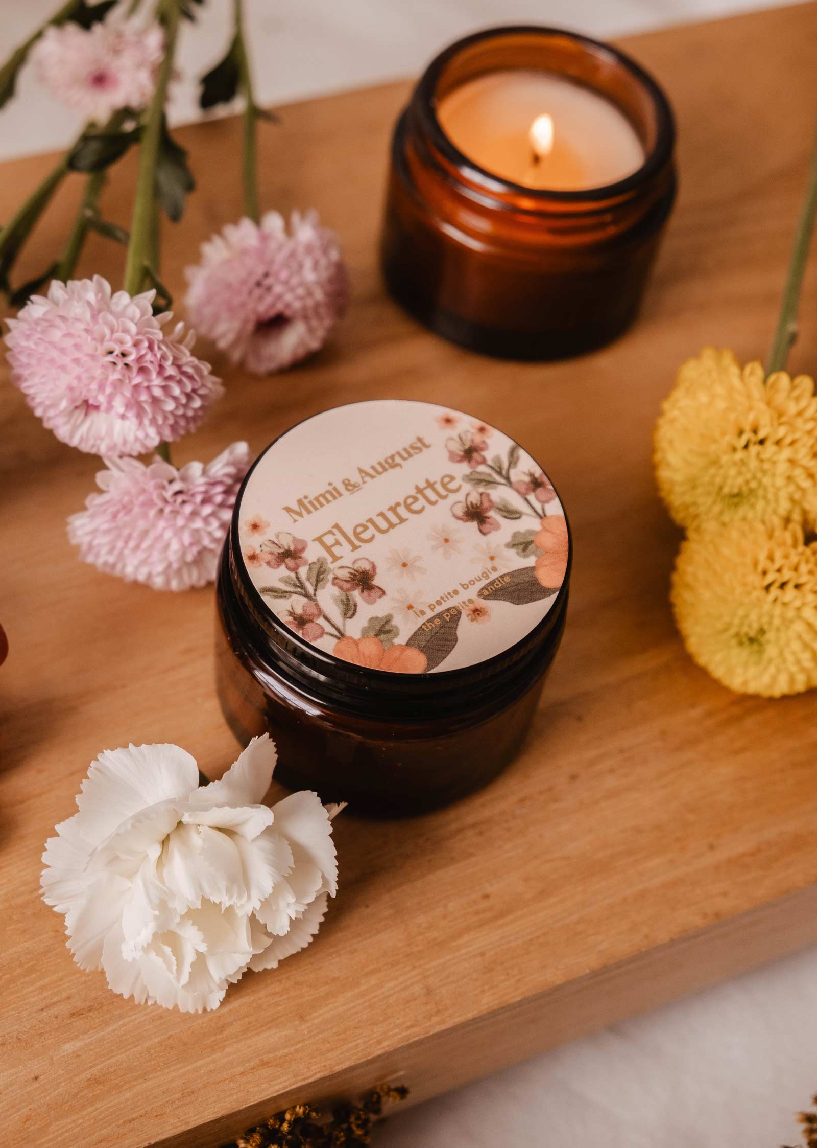 A Mimi & August "Fleurette - Reusable Candle" featuring a floral design sits on wood, surrounded by wild pink and yellow flowers. The lit candle in a brown jar fills the air with delicate bergamot and floral scents.