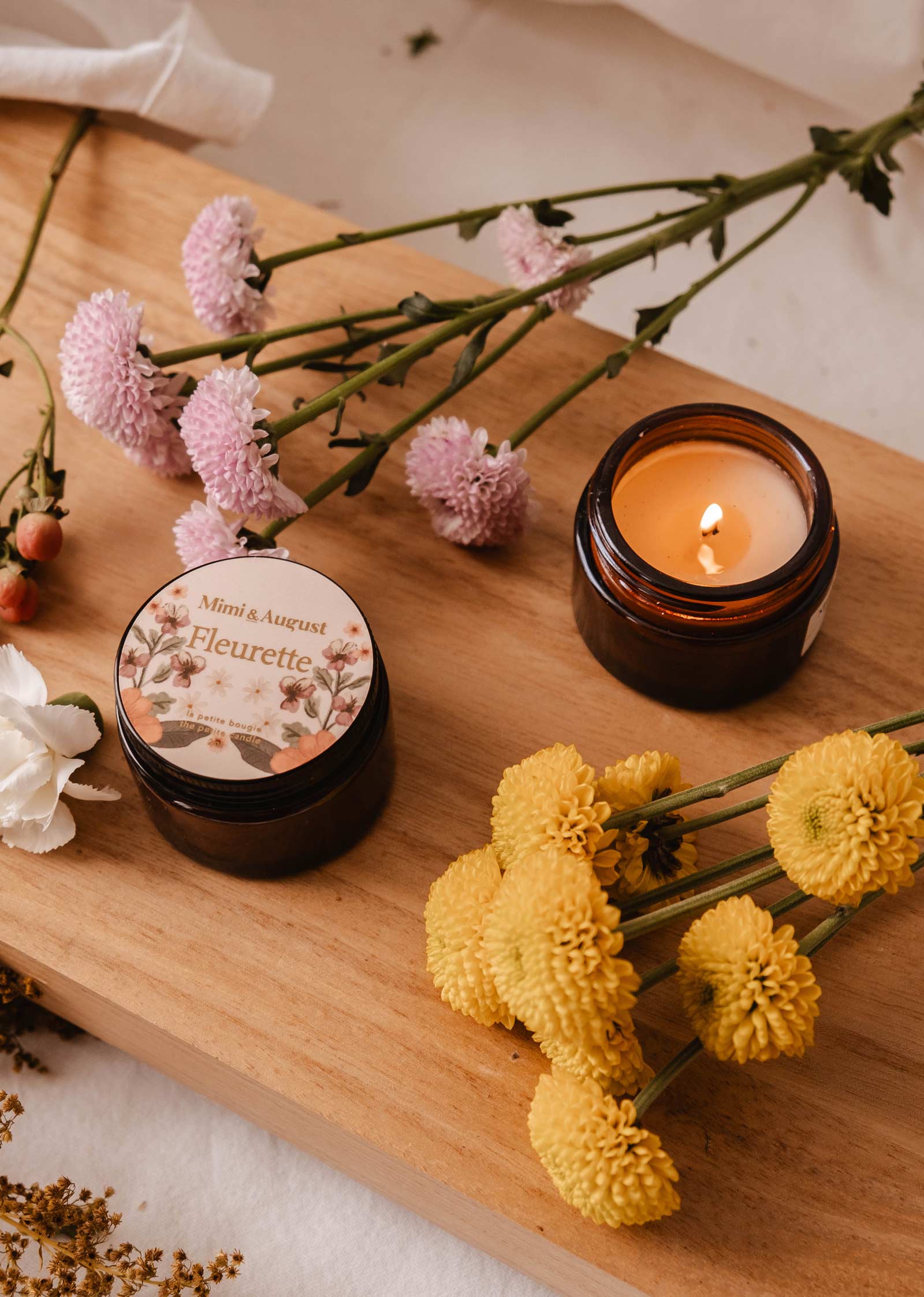 A brown jar labeled "Fleurette," a lit candle by Mimi & August, sits with another jar, both surrounded by pink and yellow wildflowers on wood. The air carries enchanting floral scents with subtle bergamot hints.