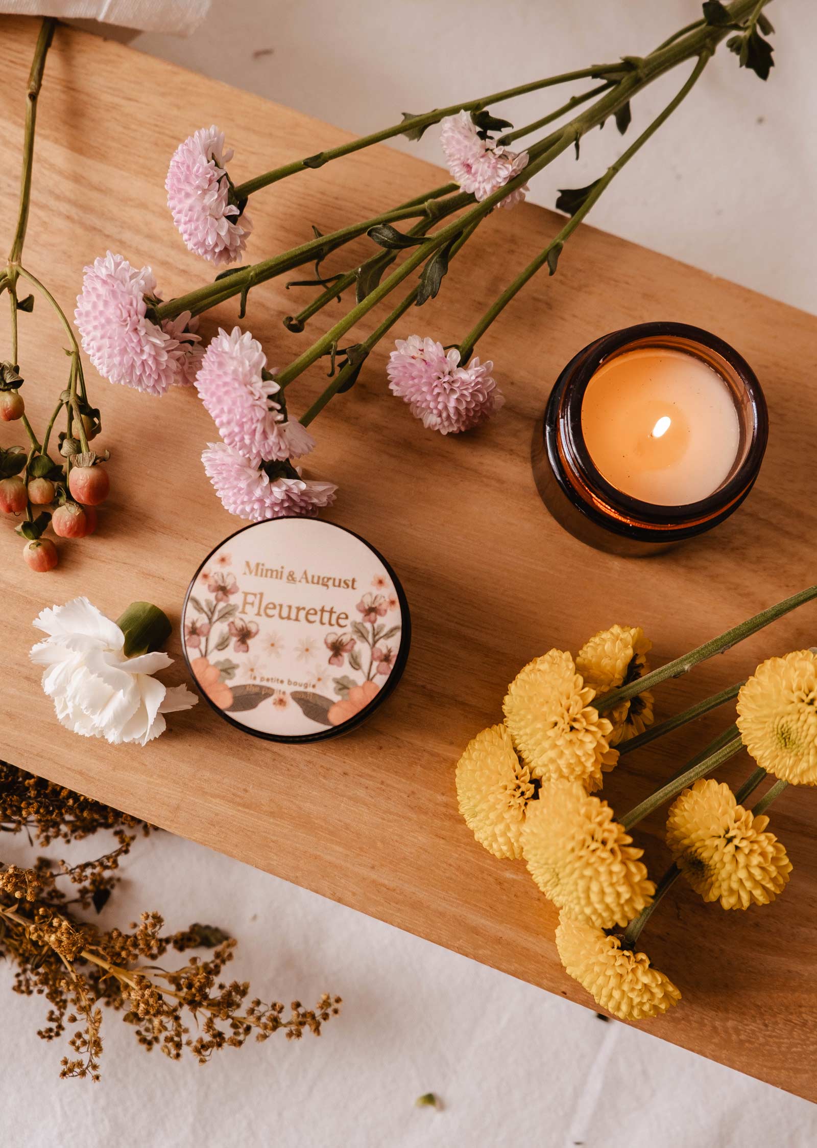 Pink and yellow flowers surround a lit Fleurette - Reusable Candle by Mimi & August on a wooden surface, releasing floral scents from its round tin.