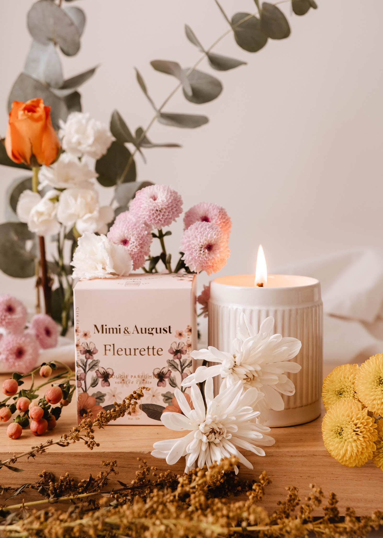 A lit Fleurette - Reusable Candle by Mimi & August sits in a floral-patterned box amidst vibrant white, pink, and yellow wildflowers on the wooden surface, with bergamot notes enhancing the scents.