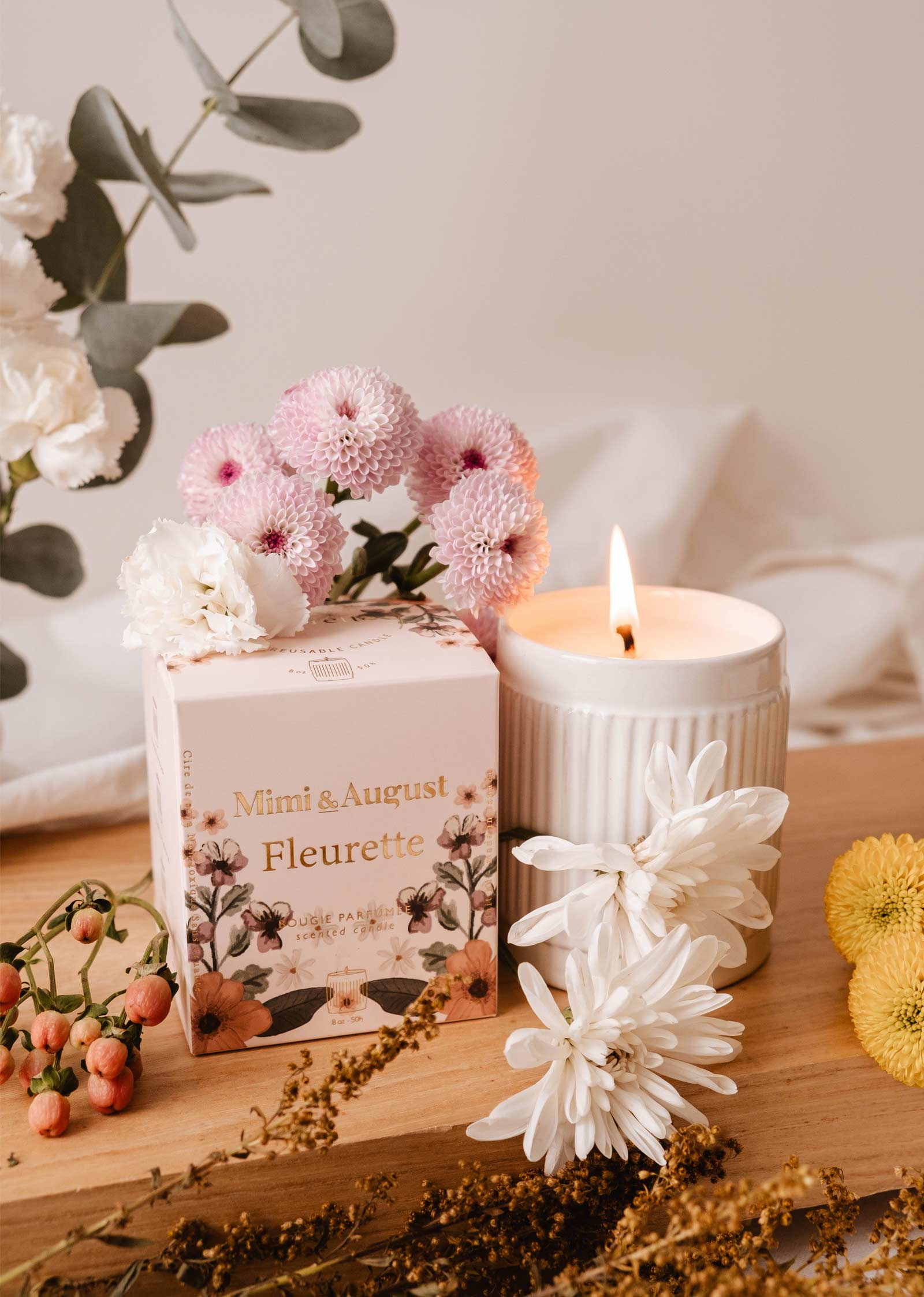 A lit "Fleurette - Reusable Candle" by Mimi & August emits lovely floral aromas amidst wildflowers on a wooden surface, near its box.