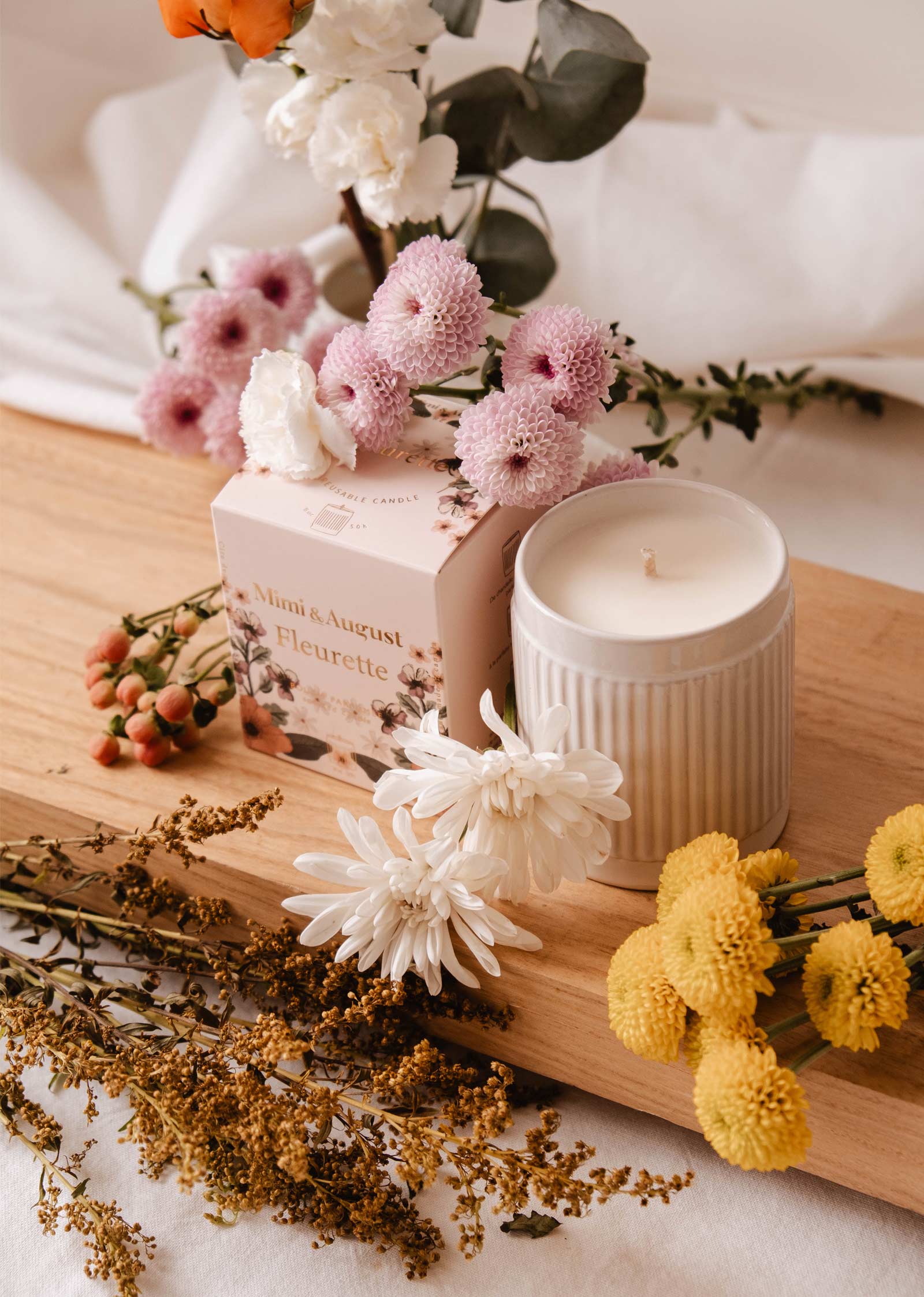 The Fleurette Reusable Candle by Mimi & August, housed in a ribbed container, is nestled among pink, white, and yellow wildflowers on a wooden surface. Its patterned box infuses the air with delicate floral scents and bergamot hints.