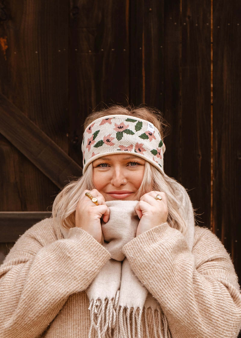 Une femme blonde tient son écharpe beige près de son visage, portant le bandeau chaud et confortable "Floral Blossom Headband" de Mimi & August. Elle est vêtue d'un pull beige et se tient debout sur un fond de bois sombre.